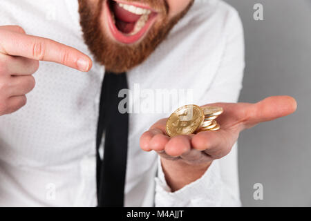 Cropped image of an excited businessman pointing finger at golden bitcoins in his arm isolated over gray background Stock Photo