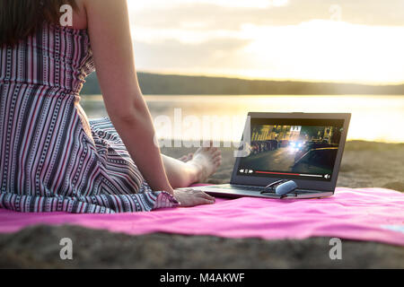 Young woman streaming a movie with laptop computer on beach at sunset. Watching film stream on imaginary online service outdoors. Video player on scre