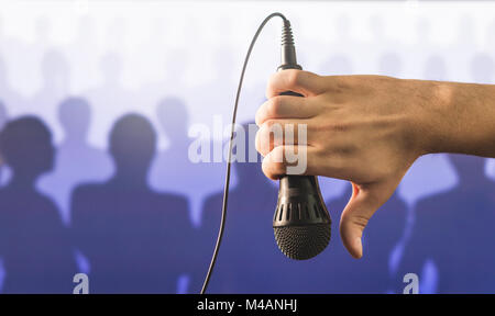 Hand holding microphone and showing thumbs down in front of a crowd of silhouette people. Failed public speaking and giving speech concept. Stock Photo