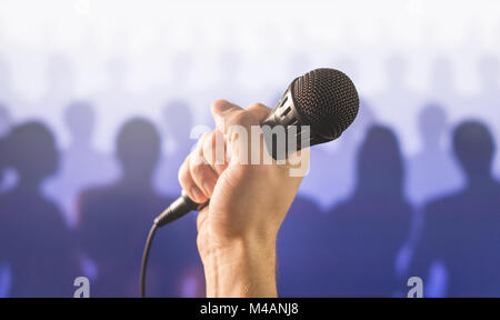 Public speaking and giving speech concept. Hand holding microphone in front of a silhouette audience and crowd of people. Singing to mic in karaoke. Stock Photo