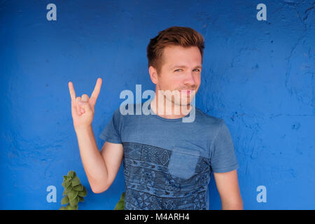 Young excited caucasian man in blue t-shirt, showing horns hand gesture with one hand and smiling. Stock Photo