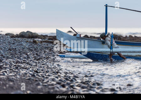 balinese fisherman returns back from the fishing and pushes his outrigger canoe (Jukung) to the gravel beach Stock Photo