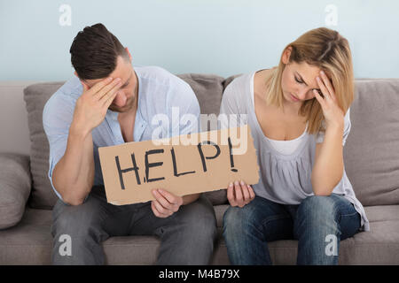 Worried Young Couple Sitting On Couch Holding Help Sign At Home Stock Photo