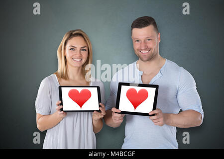 Smiling Young Couple Showing Heart On Digital Tablet Against Grey Background Stock Photo