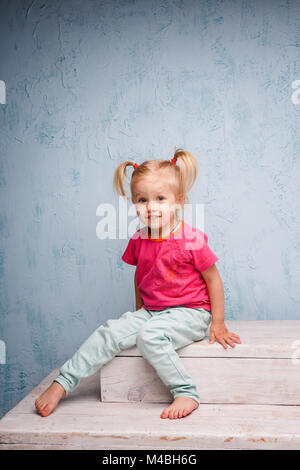 Little funny blue-eyed girl child blonde with a haircut two ponytails on her head sitting on a gossip on the background of an old textured wall in blue. Dressed in stylish bright clothes Stock Photo