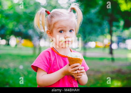 little funny girl blonde eating sweet blue ice cream in a waffle cup on a green summer background in the park. smeared her face and cheeks and laughs. Dressed in bright stylish clothes Stock Photo