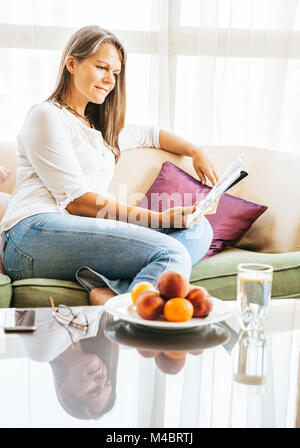 Mature woman is relaxing on a couch in her apartment with a magazine Stock Photo