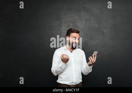 Delighted businessman in white shirt acting like winner clenching fist while using cellphone over graphite background Stock Photo