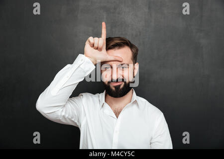 Portrait of unhappy guy 30s posing on camera and showing loser sign on his forehead over graphite background Stock Photo