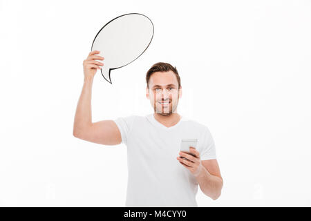 Picture of young man standing isolated over white background holding speech bubble using phone while looking camera. Stock Photo