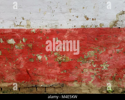 Abstract bright grunge texture background old destroyed wall of a joyful red color, on top of a white stripe, at the bottom of the wall small green pl Stock Photo