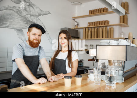 Coffee Business Concept - Positive young bearded man and beautiful attractive lady barista couple in apron looking at camera while standing at bar Counter. Stock Photo