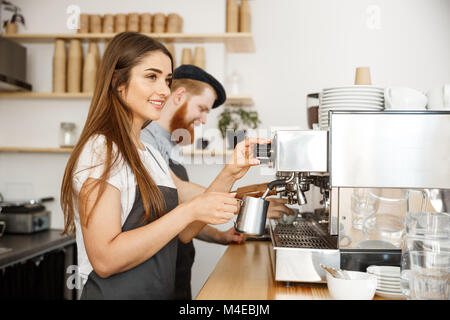 Coffee Business Concept - portrait of lady barista in apron preparing and steaming milk for coffee order with her partner while standing at cafe. Stock Photo