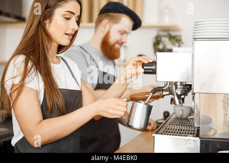 Coffee Business Concept - portrait of lady barista in apron preparing and steaming milk for coffee order with her partner while standing at cafe. Stock Photo