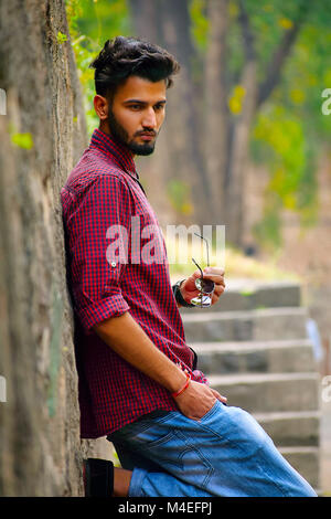 Young man with checked shirt with sun glasses in hand resting on a tree trunk, Pune, Maharashtra. Stock Photo