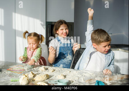 Three children prepare something from the dough. Two sisters and a brother are sitting at the kitchen table. There are various kitchen utensils on the Stock Photo