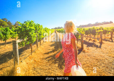 Australian vineyard. Caucasian farmer walks carefree among the rows of white grapes. Blonde female with red dress and hat getting ready for harvest. Sunset light. Margaret River, Western Australia. Stock Photo