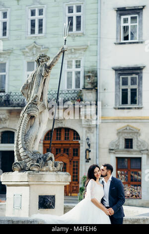 Vertical wedding portrait of the handsome groom softly kissing the gorgeous bride in the cheek while sitting on the old fountain in the city center. Stock Photo