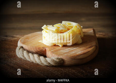 Nest of pasta tagliatelle on wooden cutting board aon rustic background. Selective focus Stock Photo