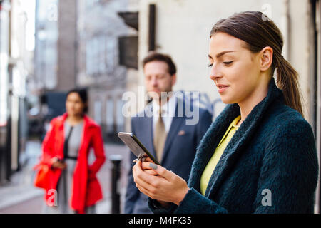 Young businesswoman is using her smartphone to book a taxi to her workplace. There is another man waiting at the taxi rank who can be seen defocussed  Stock Photo
