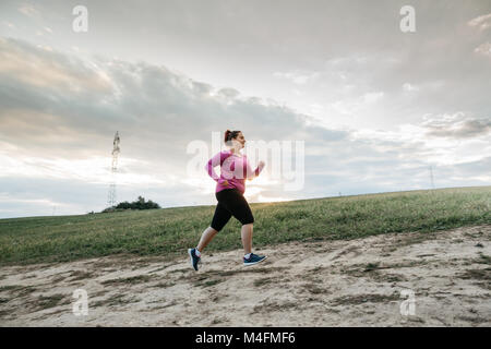 Low angle view of a plump female jogger jogging up a hill at sunset. Stock Photo