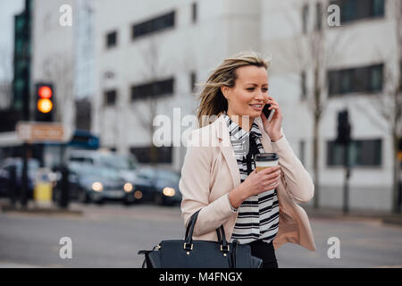 Businesswoman is walking to work in the city. She is holding a coffee cup and is talking on the phone. Stock Photo