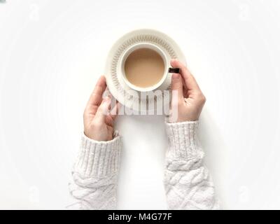 Looking down from above onto a white background with a girl's arms holding a cup of tea in a cup and saucer whilst wearing a cosy and warm swaeater. Stock Photo