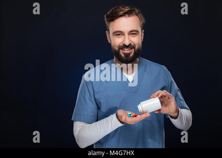 Portrait of a smiling male doctor dressed in uniform getting pills from a bottle isolated over dark background Stock Photo