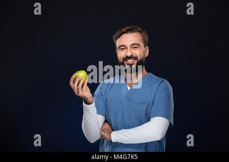 Portrait of a happy male doctor dressed in uniform holding green apple and looking at camera isolated over dark background Stock Photo