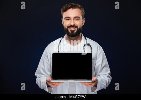 Portrait of a confident male doctor dressed in uniform with stethoscope showing blank screen laptop computer isolated over dark background Stock Photo