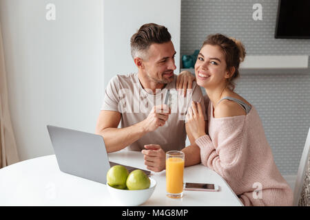 Portrait of an attractive young couple shopping online with laptop computer while sitting at the table in a kitchen at home, man holding credit card Stock Photo
