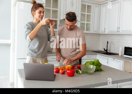 Portrait of a young loving couple cooking salad together with laptop computer while standing on a kitchen at home, woman taking picture of food on a t Stock Photo