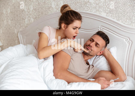 Lovely couple lying together in bed while man using his smartphone and shocked woman looking on it at home Stock Photo