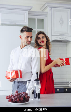 Pretty smiling lovers, handsome man and young lady in red dress, holding present to each other at home on celebrating Stock Photo