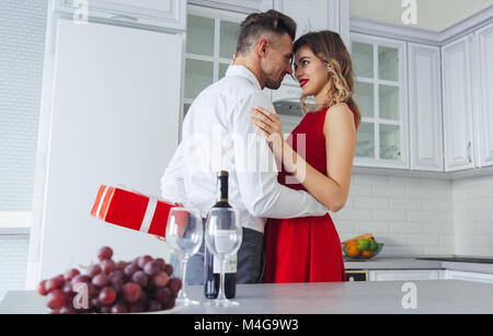 Smiling delightful lady in pretty red dress hugging her man while he hiding gift for her on celebration Stock Photo