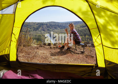 Young blonde woman tourists in camp on cliff over river and forest landscape. View from camping tent entrance. Stock Photo