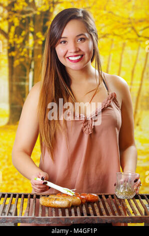 Close up of beautiful young smiling woman using a long white onion on grilling sausages on barbecue grill . BBQ in the garden. Bavarian sausages in a blurred background Stock Photo