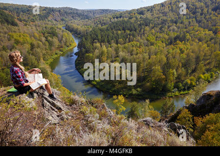 Young blonde woman tourist  sitting on a cliff with map in hands on background of siberian autumn landscape with the river Berd. Russia, Siberia, Sala Stock Photo