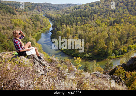 Young blonde woman tourist  sitting on a cliff with map in hands on background of siberian autumn landscape with the river Berd. Russia, Siberia, Sala Stock Photo