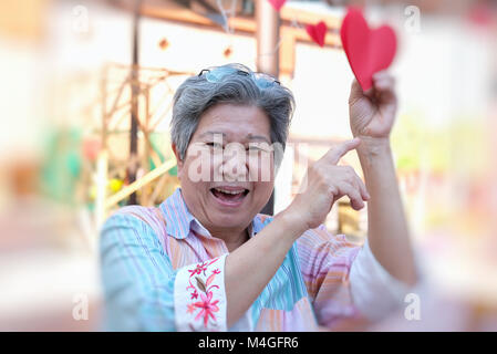asian elder woman holding red heart paper & smiling at camera. happy elderly female relaxing outdoors. mature lifestyle concept Stock Photo