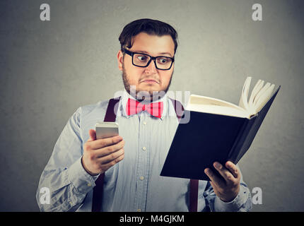 Young chubby man holding printed book and smartphone looking extremely puzzled. Stock Photo