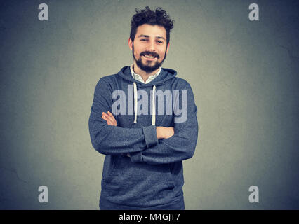 Young hipster man in gray hoodie standing with hands crossed smiling happily at camera. Stock Photo