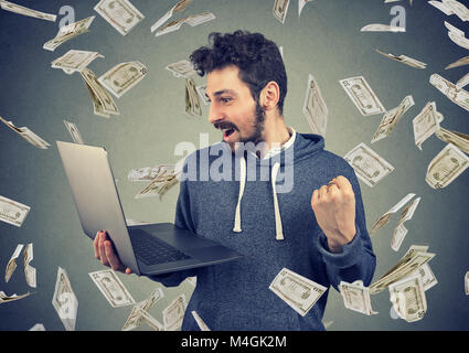 Successful young man using laptop building online business making money dollar bills cash falling down. Money rain. Beginner IT entrepreneur success e Stock Photo