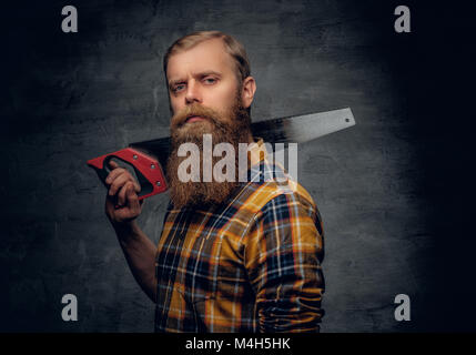 A bearded carpenter man dressed in a plaid shirt holds handsaw on a shoulder. Grey vignette background. Stock Photo