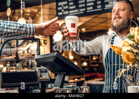 Positive bearded barista male selling coffee to a consumer in a coffee shop. Stock Photo