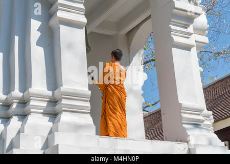 CHIANG MAI THAILAND - JANUARY 29 2018; Buddhist Monk in orange robes stands  on high level of white ornate bell tower ringing bell Stock Photo
