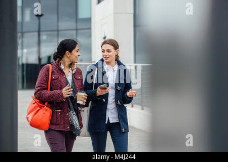 Businesswomen are walking to work together through the city. They are talking and one is holding a coffee while the other is holding a smartphone. Stock Photo