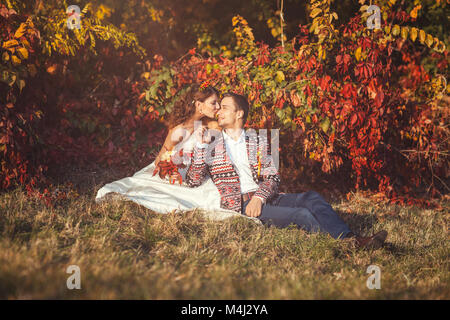 Newlyweds embracing sitting on the grass near the tree in the autumn forest, bride kisses groom Stock Photo