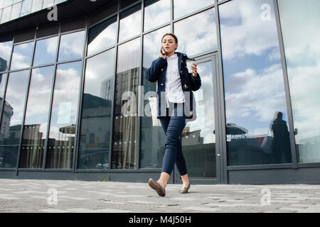Young businesswoman is leaving her office building with a cup of tea. She is talking on the phone to someone as she walks. Stock Photo