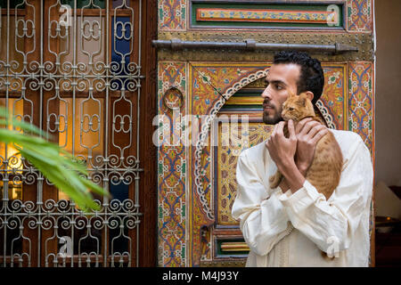 Young muslim man in traditional clothing holding a yellow cat in traditional Moroccan ambient in front of a decorated wall Stock Photo